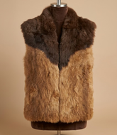 A fur vest displayed on a hanger stand inside a workshop or showroom. The vest features a dark brown collar and upper section, transitioning into a warm tan and light brown textured fur body.