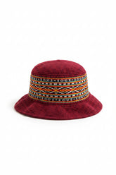 A bright pinkish-red crocheted bucket hat with a wide, colorful geometric patterned band around the crown, resting on a gray stone tile floor.