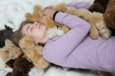 A girl lying on a pearl alpaca rug surrounded by plush teddy bears, wearing a lavender t-shirt embracing the teddy bears.
