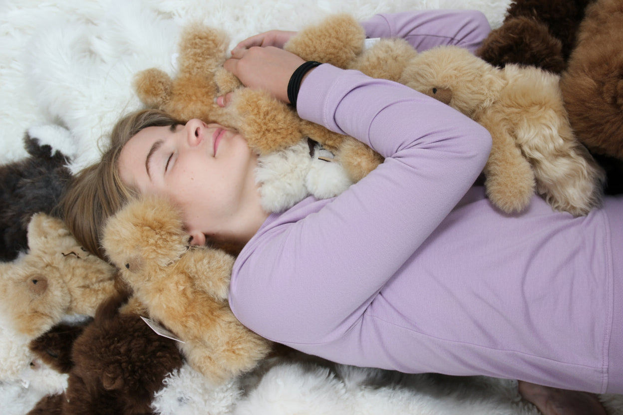 A girl lying on a pearl alpaca rug surrounded by plush teddy bears, wearing a lavender t-shirt embracing the teddy bears.