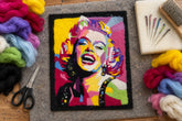 A framed pop-art style portrait print of Marilyn Monroe, featuring a vibrant multicolor design with a gold-toned frame, displayed on a white sheet of paper over a wood-grain surface.