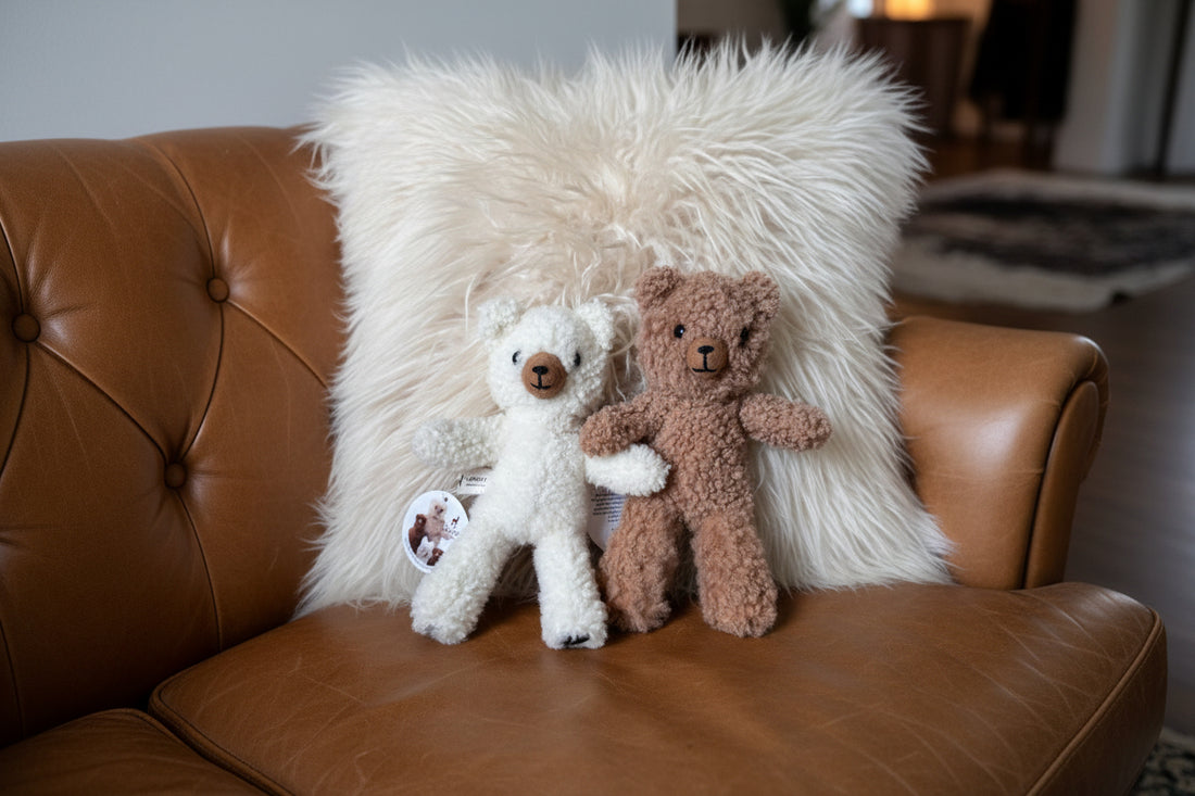 Two teddy bears, one cream white and one medium brown, made from curly textured fabric, posed together on a striped armchair in front of a fluffy cream throw pillow.
