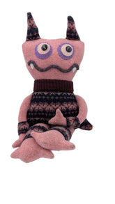A pink knitted monster plush toy with dark brown and black patterned horns, a dark brown knit turtleneck, and a dark patterned sweater, sitting on a white surface against a white background.