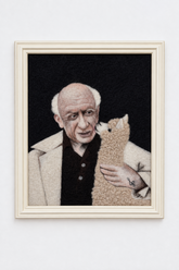A framed portrait artwork leaning against a white wall on a wooden floor. The artwork shows a bald elderly man in a light coat holding a beige alpaca-like animal against a dark background.