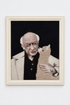A framed portrait artwork leaning against a white wall on a wooden floor. The artwork shows a bald elderly man in a light coat holding a beige alpaca-like animal against a dark background.