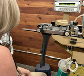 A person seated beside a large industrial sewing or cutting machine in a workshop, with fabric moving through the machine and a digital control panel visible above.