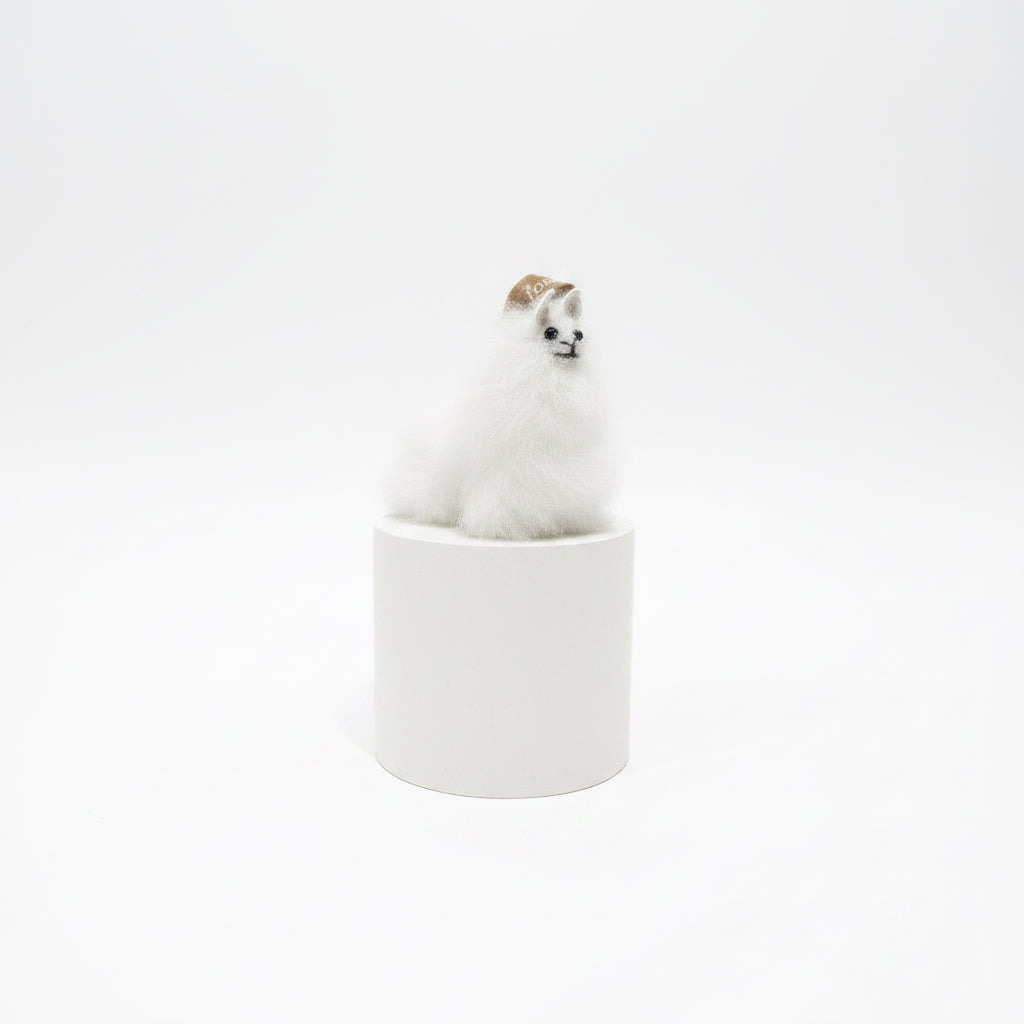 A small white fluffy animal figurine, resembling a llama or alpaca, wearing a tan hat, sitting on top of a striped spool-shaped base against a textured beige wall and dark brown tabletop.