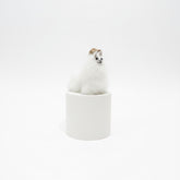 A small white fluffy animal figurine, resembling a llama or alpaca, wearing a tan hat, sitting on top of a striped spool-shaped base against a textured beige wall and dark brown tabletop.