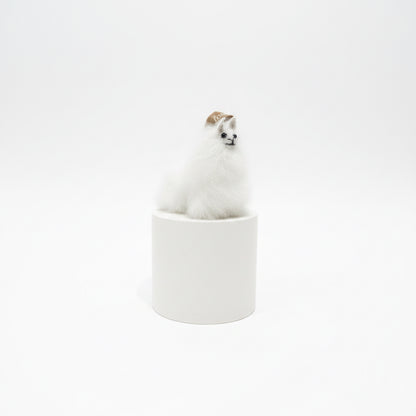 A small white fluffy animal figurine, resembling a llama or alpaca, wearing a tan hat, sitting on top of a striped spool-shaped base against a textured beige wall and dark brown tabletop.