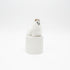 A small white fluffy animal figurine, resembling a llama or alpaca, wearing a tan hat, sitting on top of a striped spool-shaped base against a textured beige wall and dark brown tabletop.