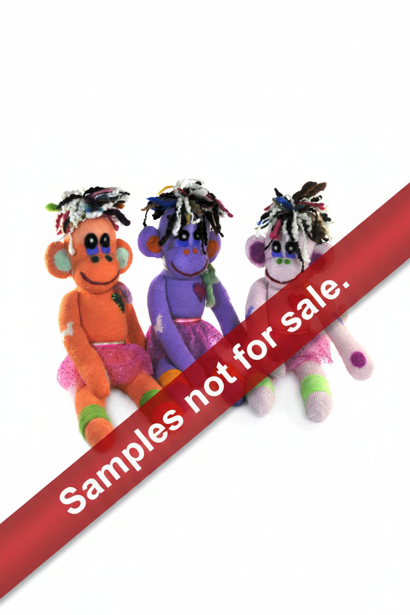 Three handcrafted sock monkey dolls with orange, purple, and pink coloring, each with a unique facial expression and decorative elements such as heart-shaped badges.