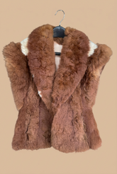 A brown fur vest with a wide collar and cream lining, hanging on a black hanger in a workshop setting.