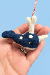 A hand holding a small felted wool whale ornament. The whale is dark blue with white spots and white flippers, featuring a pink braided hanging loop.