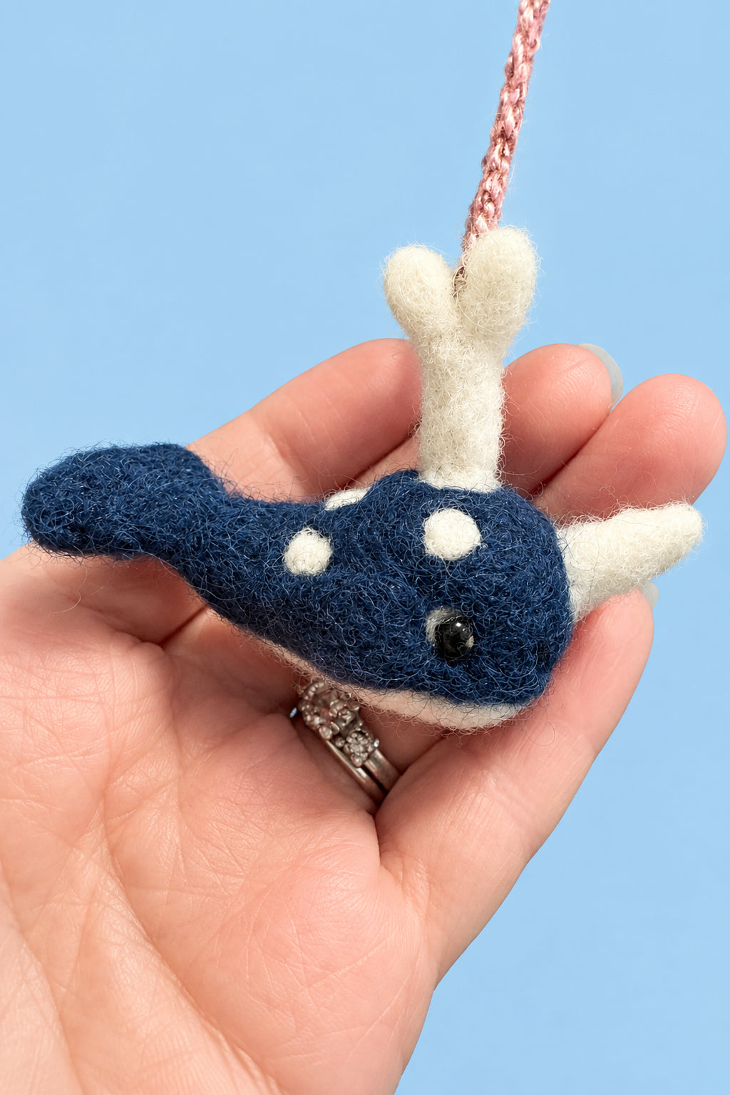 A hand holding a small felted wool whale ornament. The whale is dark blue with white spots and white flippers, featuring a pink braided hanging loop.